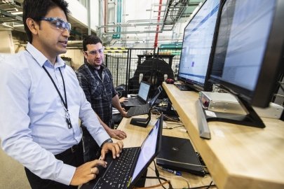 Two researchers standing in front of two large computer screens and two laptops. One of the researchers is moving the mouse on one of the laptops.