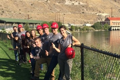 A group of students standing near a body of water and a hydropower dam