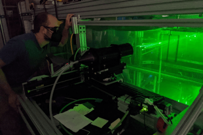 Person with eye protection looking at green beams of light in a contained clear box.