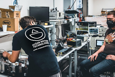 Two people working on machinery on top of a desk. The back of one person’s shirt reads “OCEAN MOTION TECHNOLOGIES.” 