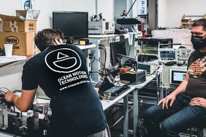 Two people working on machinery on top of a desk. The back of one person’s shirt reads “OCEAN MOTION TECHNOLOGIES.”