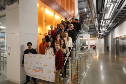 A group of students standing on an indoor staircase. Those in the front hold a banner patterned with cartoon cowboy hats and wind turbines overlain with the Cyclone Cowboys logo.