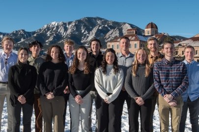 Two rows of students standing in snow in front of brick buildings and mountains.