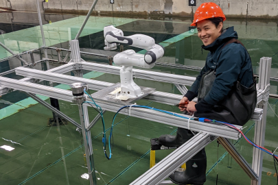 A student wearing a hard hat and waterproof attire sits on a structural beam above a tank of water. A robotic arm is attached to the beam next to him and a surfboard and offshore wind turbine tower float in the water below.