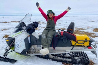 Serina Wesen (a white woman in snow overalls, snow boots, and snow gloves) sitting on a snow machine full of gear on the tundra with melting snow