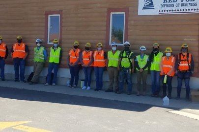 13 students wearing construction hats and vest stand in the shade along a building.