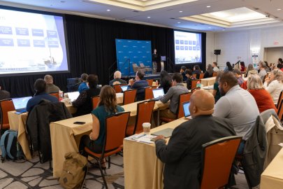 An audience watches a presentation in a hotel ballroom.