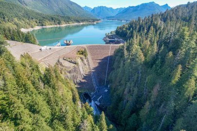 A large dam and reservoir, nestled among forests and mountains, as seen from above