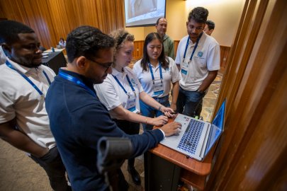Several people stand around a laptop, looking at it while two people work on it
