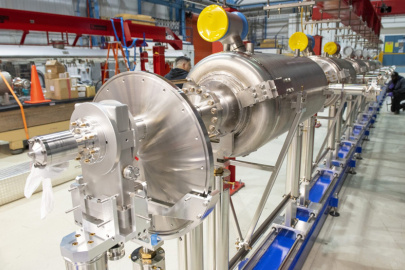 A long view of the fully assembled prototype high-beta 650-megahertz cryomodule shows a glossy steel horizontal structure that is large and heavy. It is in a lab area where it is being built out. Four of these will make up the final stage in Fermilab’s new linear accelerator.