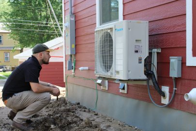 A crouching person inspects an air-source heat pump mounted to the side of a home.