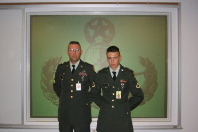 Two men in military uniforms stand and pose for a picture, they stand in front of a green and white wall with a black logo printed it