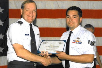 Two men in military uniforms shake hands and hold a certificate while standing in front of an American flag