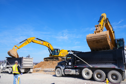 Two work machine vehicles scoop dirt into the back of two large trucks, workers stand around in yellow protective vests