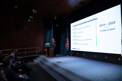 A woman standing at a podium giving a presentation to a crowd. There is a screen to her right with statistics displayed.