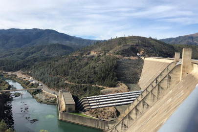 Side view of a large hydropower dam with the outflowing river and tree lined valley in the background