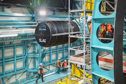 The giant telephoto lens of the LSST Camera as it is being placed on the Rubin Observatory, surrounded by scaffolding. People are below it in orange safety gear.