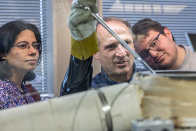 Three scientists watch as the center one holds a tool with a gloved hand.