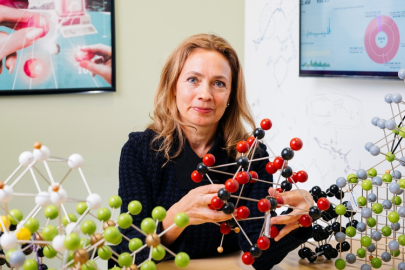 Kristin Persson (a woman in a black shirt) sitting at a desk in an office. She is holding a complex molecular structure and there are several molecular structures on the table in front of her. 
