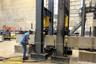 man in blue shirt and white helmet stands next to horizontal column of gray concrete inside test facility