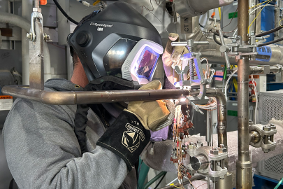 A worker wearing a welding mask welds a pipe onto the new cold trap device at the METL facility.