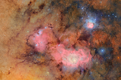 A tapestry of glowing tan and pink gas clouds with dark dust lanes. In the upper right, the Trifid Nebula resembles a flower with pinkish gas petals surrounded by blue gas, and streaked with dark, finger-like veins of dust. To the lower left, the much larger Lagoon Nebula stretches like a sea of magenta gas, with blue knotted clumps throughout. Both nebulae are embedded in a tan backdrop of gas that is brighter on the left than the right, etched with tendrils of dust and sprinkled with stars.