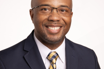 A portrait of a man in glasses and a black suit with a yellow and black tie