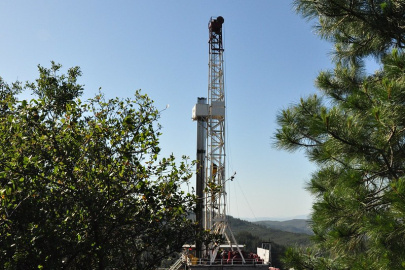 Photo of a drill rig between trees in the foreground, with rolling hills extending into the background.