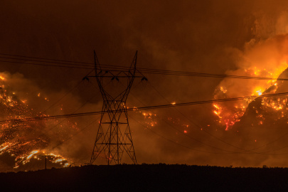 Transmission towers and power lines silhouetted against a large wildfire burning across a hillside at night.