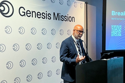 A man in a suit speaking at a podium with a presentation to his right and a white background with the words "Genesis Mission" written at the top 