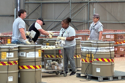 A group of employees in grey shirts move large light brown buckets with red and yellow striped tape around them inside a facility building