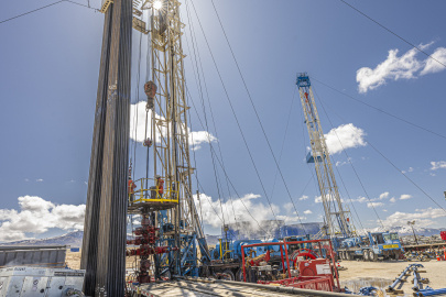 Close-up photo of a drill rig stretching up into a blue sky with the sun shining right behind the derrick (i.e. the tower).