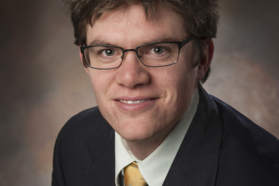 Headshot of a white man with brown hair and glasses in a black suit with gold tie