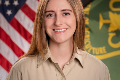 A professional headshot of a Forest Service fellow in a park ranger outfit with an American flag and Forest Service flag in the background