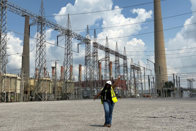 A woman in a white safety hat and yellow safety vest poses for a photo in front of tall electrical towers