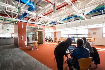 Photo of a large warehouse-looking room with three people gathered around a laptop on a small table.