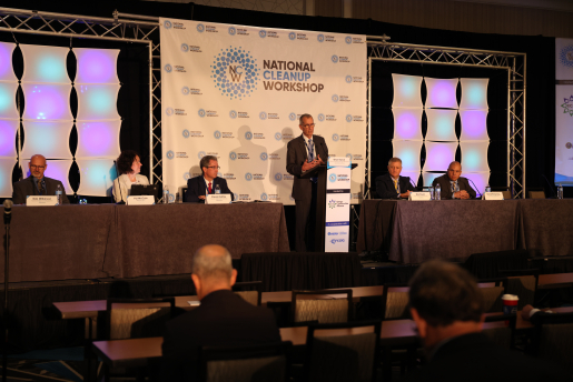 Three people sit at a table and two people sit at another long table with a man at a podium in between them.