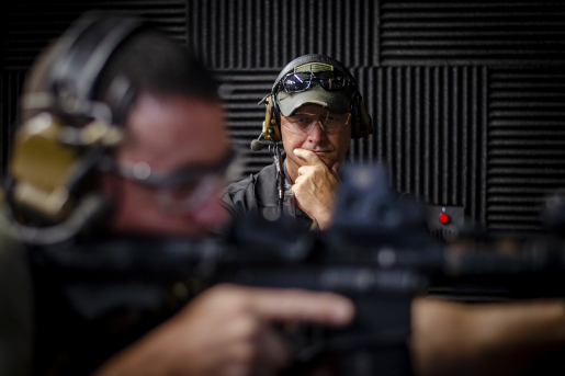 Person aiming down the sites of a rifle with person observing in the background.