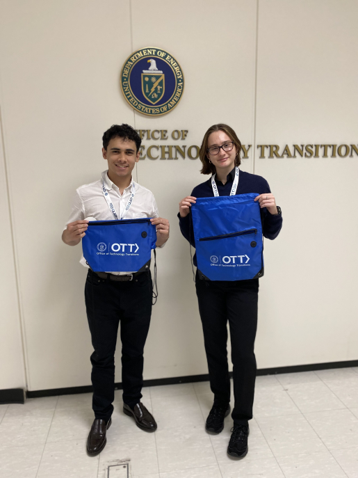 Eduardo Andujar (left) and Anna Hylsop (right) standing in front of an Office of Technology Transitions sign holding blue, OTT-branded bags