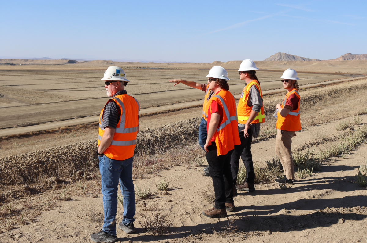 A group of people in white hard hats and orange safety vests stand in an open field with mountains in the distance behind them