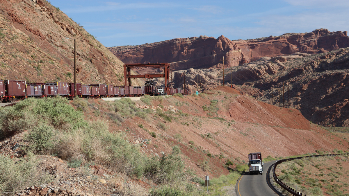 A long train on tracks above a road with a tractor trailer truck driving on it 