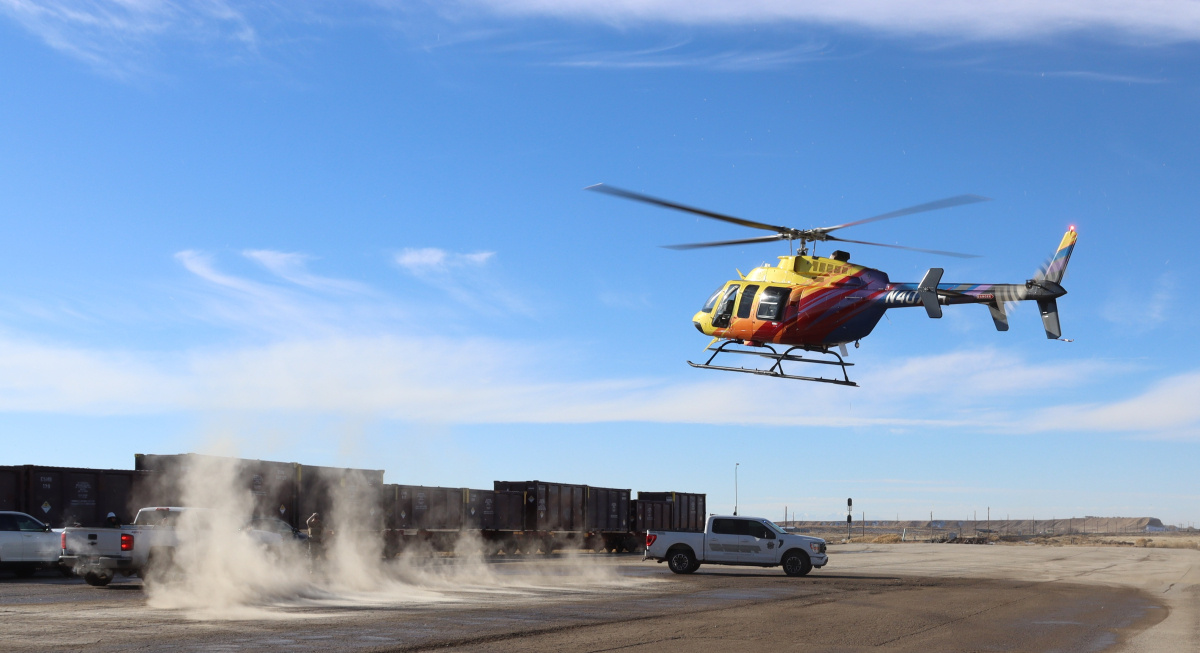 A yellow, orange, and red helicopter lands in an open space with trucks and vehicles parked nearby