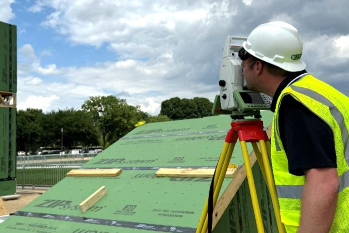Man in a hard hat and reflective vest standing near a construction site where others are working on the side of a building.