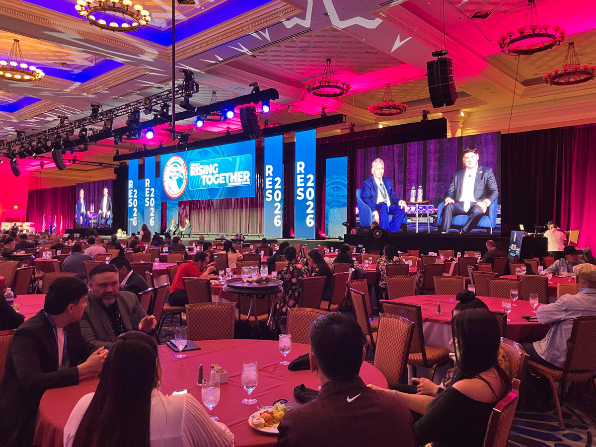 A large ballroom with people seated at round tables, watching two people sitting and speaking on stage and on a big screen.