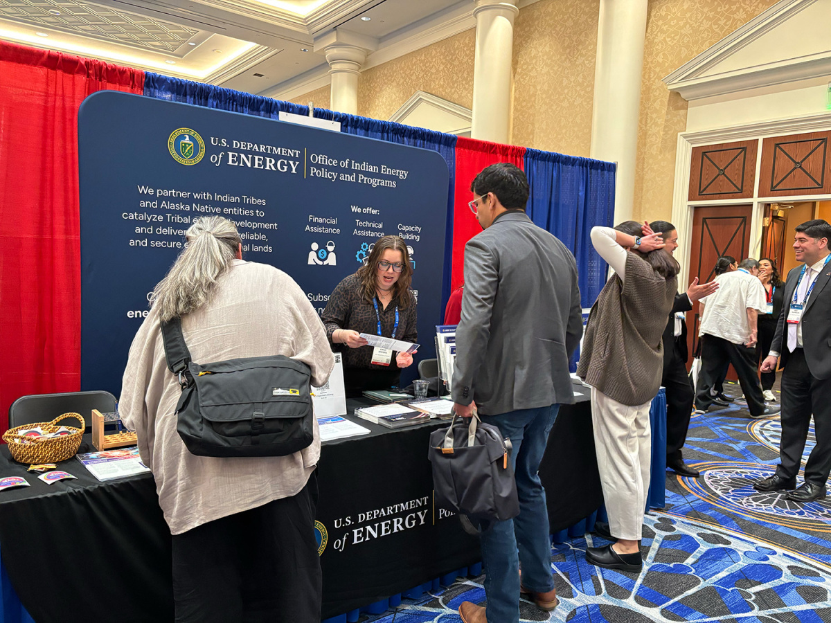 People talking at a conference booth with a large banner behind them about the Office of Indian Energy.
