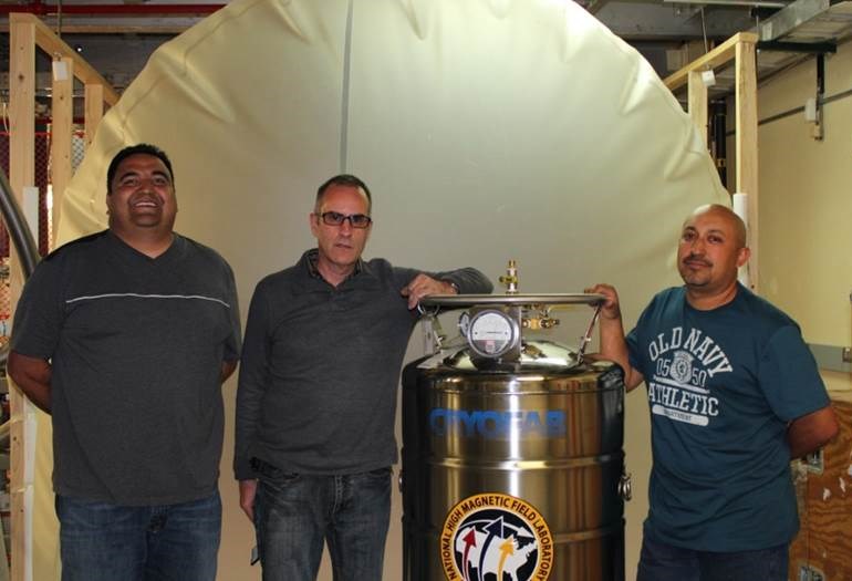 Photo of three men standing next to a stainless steel canister in a laboratory.