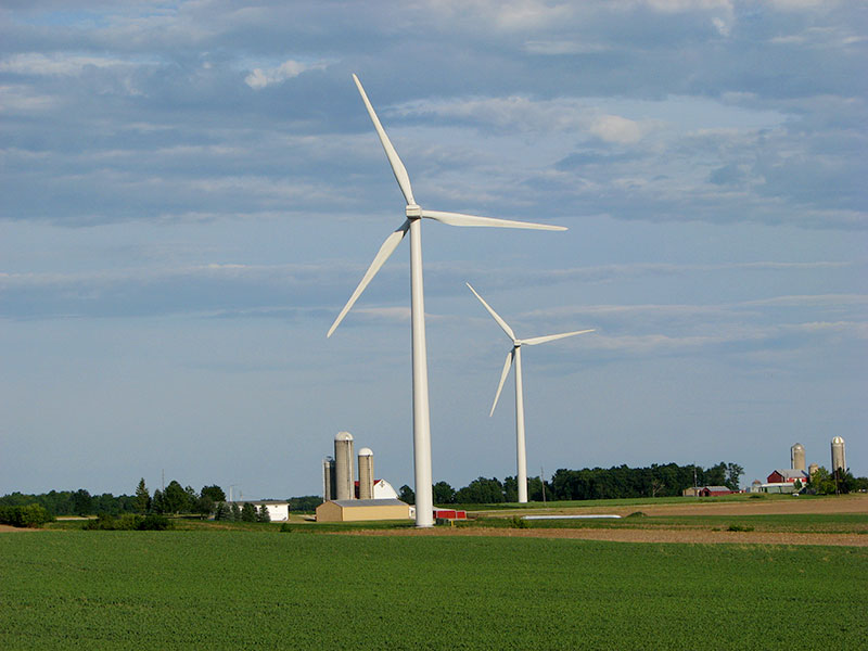 Multi-story farm buildings, grain silos, and trees look small compared to the two multimegawatt wind turbines.