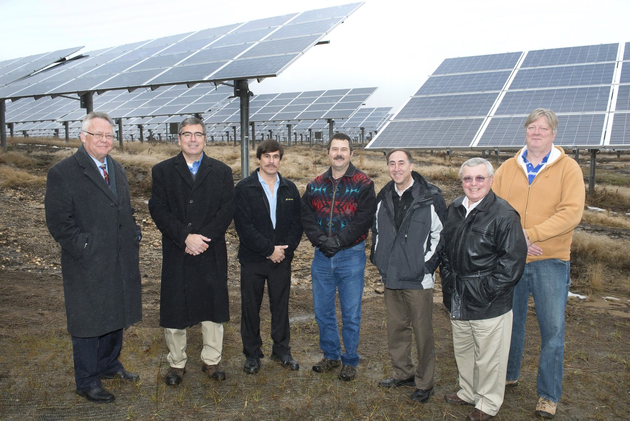 Photo of a group of seven men standing in a semi-circle in front of a field of large solar panels