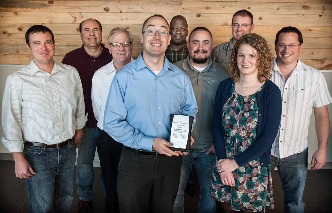 Photo of eight men and one woman gathered together. The man in the front center of the image holds an award.