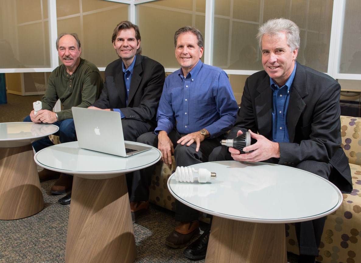 Four men sit behind three small tables; a lap top is on the table in front of the third man.